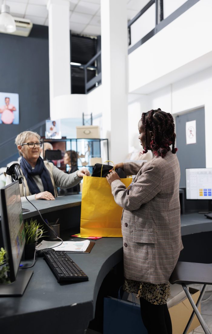 African american employee preparing bag with purchase for elderly client in modern boutique. Fashionable woman buying stylish casual wear in store, making electronic payment at pos terminal.