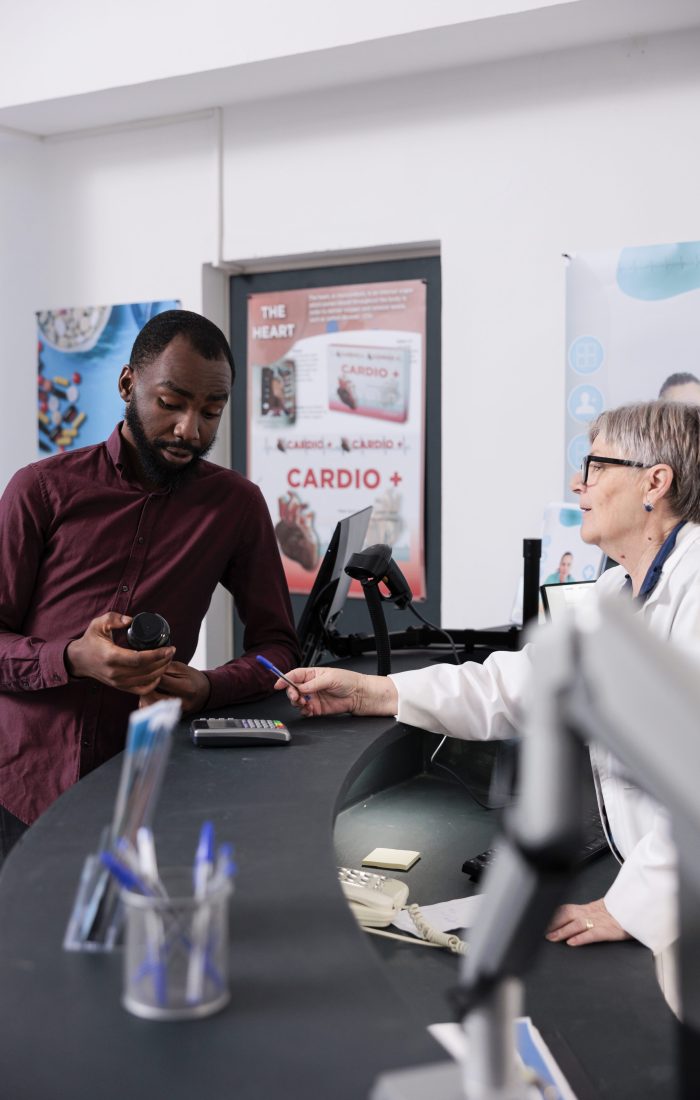 Senior drugstore worker standing at pharmacy counter helping client with pharmaceutics treatment after checking medical prescription, discussing pills leaflet. Health care service and support