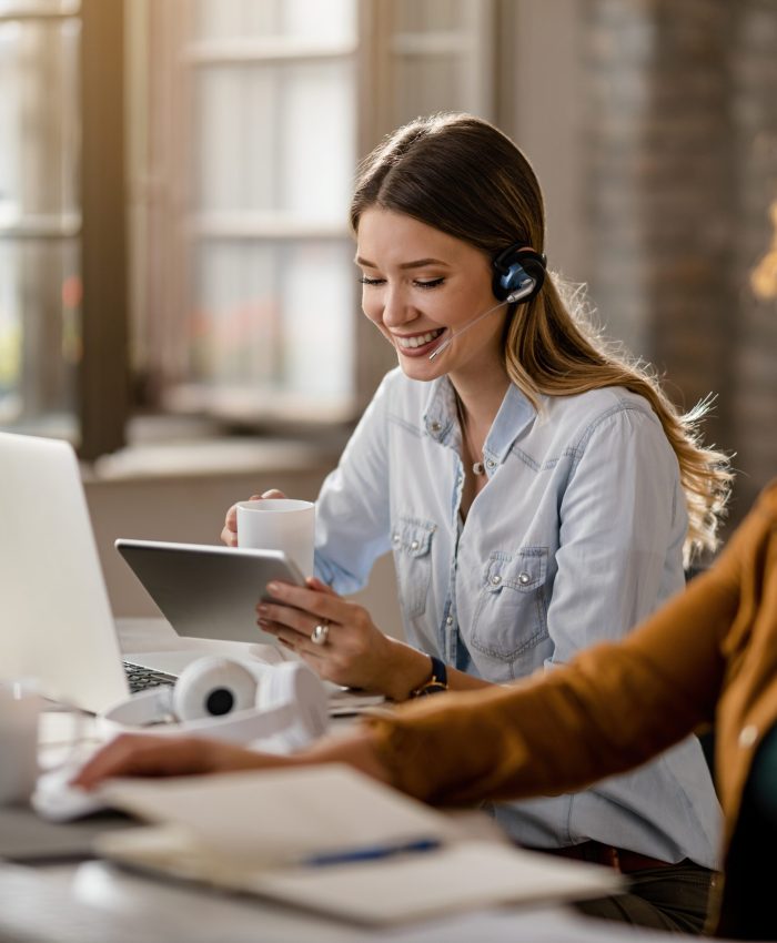 Happy female entrepreneur with headset drinking coffee while surfing the net on touchpad in the office.