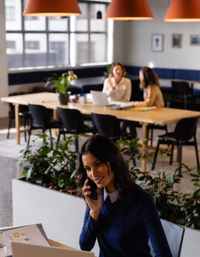 Happy middle eastern casual businesswoman using laptop and talking on smartphone at desk in office. Casual office, business, lifestyle, communication and work, unaltered.