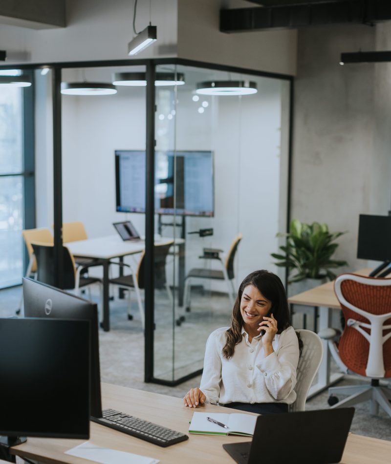 Pretty young business woman using mobile phone while working on laptop in the modern office